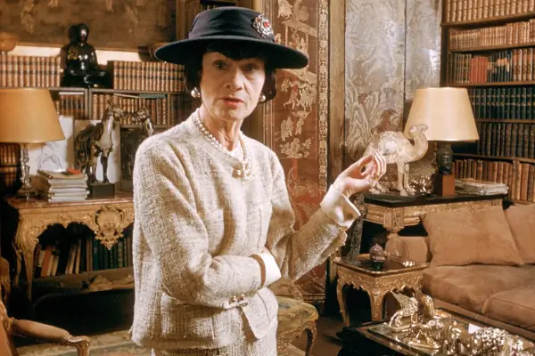 Coco Chanel em seu apartamento em Paris, vestindo tailleur de tweed com pérolas e chapéu elegante.