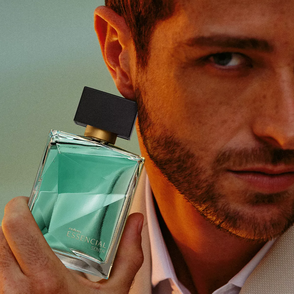 Homem com camisa clara e jaqueta marrom segura próximo ao rosto um frasco de perfume retangular com tom esverdeado e tampa preta. O frasco é o perfume da Natura Essencial Sentir masculino. A imagem destaca o produto com iluminação suave, transmitindo sofisticação e masculinidade.